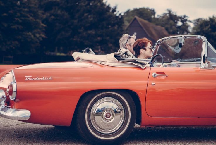An elderly couple driving in their orange classic car